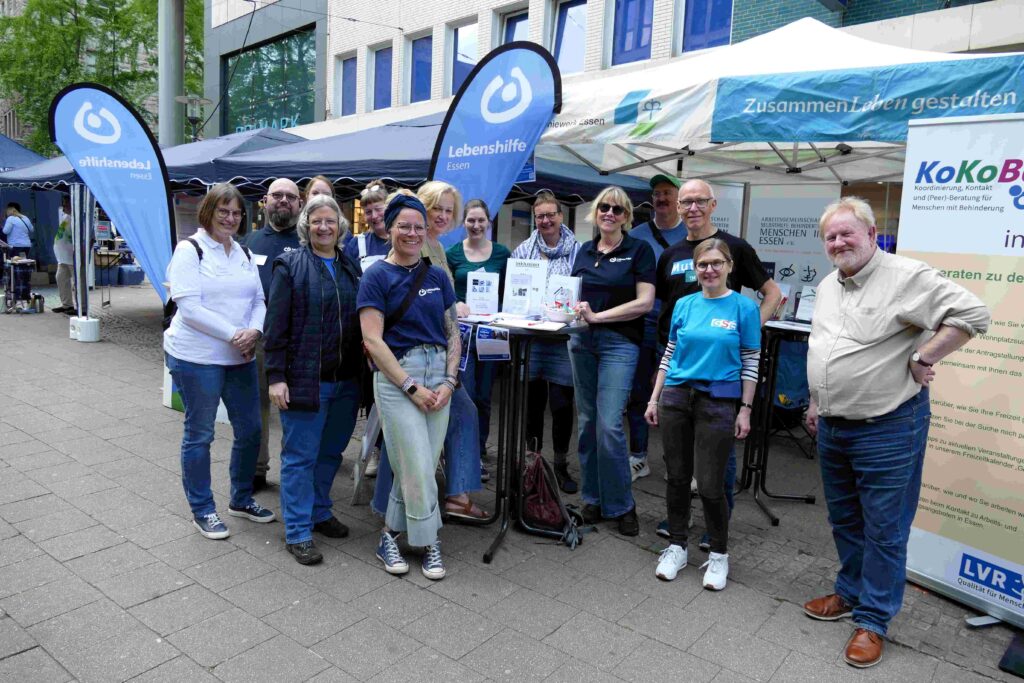 Gruppenfoto bei einer inklusiven Informationsveranstaltung in der Essener Innenstadt. Rund ein Dutzend Menschen unterschiedlichen Alters stehen vor mehreren Pavillons und Infoständen zusammen. Zwei große blaue Fahnen der Lebenshilfe Essen rahmen die Gruppe ein. Auf den Zeltdächern stehen Slogans wie „ZusammenLeben gestalten“. Einige Personen halten Informationsmaterialien in den Händen, andere lehnen entspannt an Stehtischen. Die Atmosphäre wirkt offen, freundlich und gemeinschaftlich. Im Hintergrund sind weitere Stände und die Fassade eines modernen Geschäftsgebäudes zu sehen.