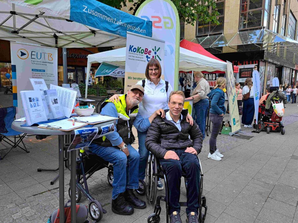 Drei Personen posieren an einem Informationsstand unter einem weißen Pavillon. Zwei Männer sitzen in Rollstühlen beziehungsweise Mobilitätshilfen, eine Frau steht lächelnd hinter ihnen und legt beiden freundschaftlich die Hände auf die Schultern. Neben ihnen stehen Infomaterialien und Aufsteller der EUTB und KoKoBe. Im Hintergrund verläuft eine belebte Fußgängerzone mit weiteren Zelten, Besucherinnen und Besuchern sowie Menschen mit unterschiedlichen Mobilitätshilfen.