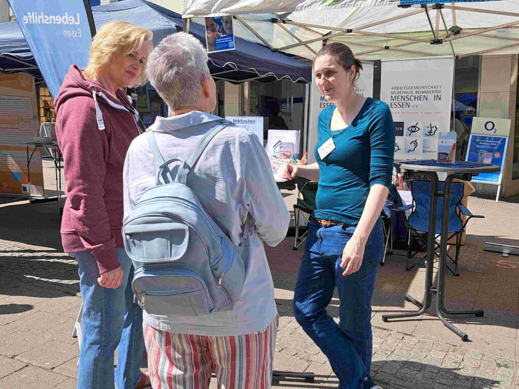 Eine junge Frau informiert zwei Besucherinnen an einem Stehtisch vor einem Pavillon der „Arbeitsgemeinschaft Selbsthilfe behinderter Menschen in Essen“. Die Beraterin trägt ein Namensschild und hält einen Stift in der Hand. Die beiden Frauen stehen ihr aufmerksam gegenüber, eine davon mit hellem Rucksack. Sonnenlicht fällt auf die Szene und erzeugt eine freundliche, sommerliche Stimmung. Im Hintergrund sind weitere Informationsstände und Logos verschiedener Organisationen sichtbar.