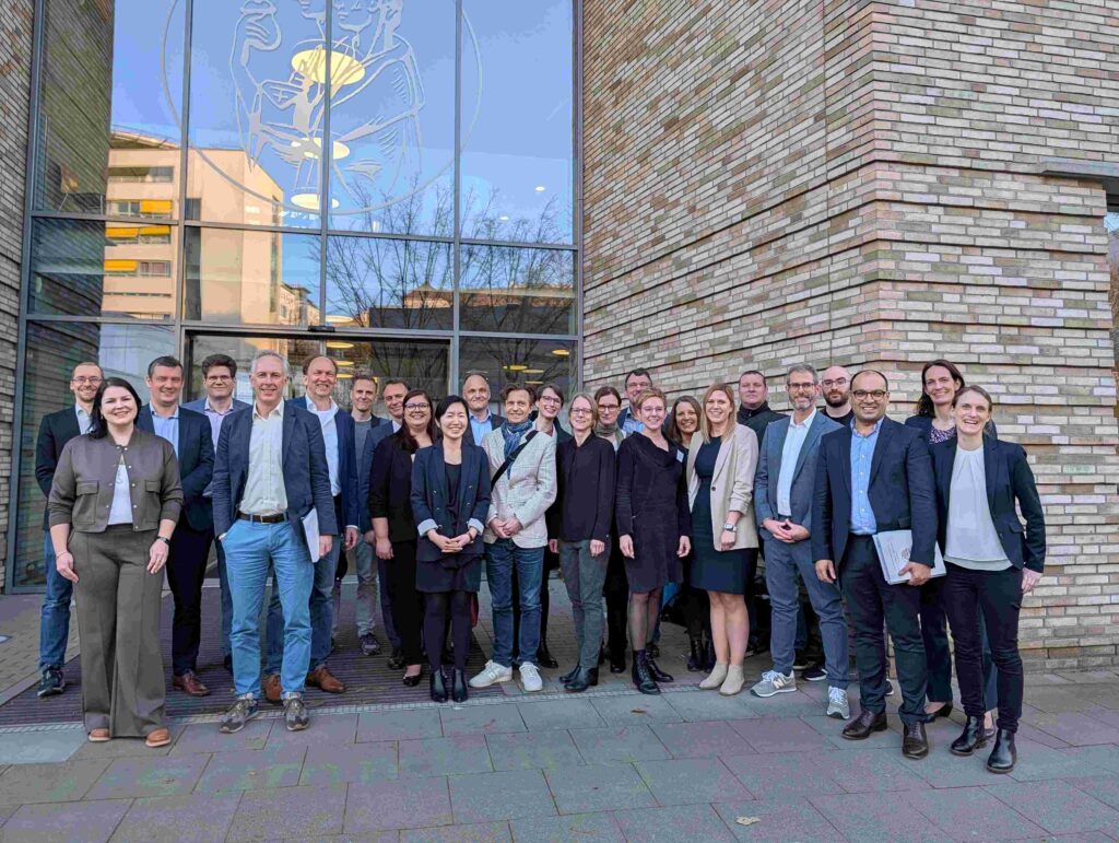 Vor einem modernen Backsteingebäude mit großer Glasfront steht eine Gruppe von rund 20 Personen für ein gemeinsames Foto. Die Teilnehmenden tragen überwiegend Business- und Smart-Casual-Kleidung und blicken lächelnd in die Kamera. Im Hintergrund spiegelt die Glasfassade den Himmel und umliegende Gebäude wider; im oberen Bereich ist eine stilisierte medizinische Darstellung in das Glas eingelassen. Die Gruppe ist locker angeordnet, einige Personen stehen leicht versetzt in mehreren Reihen. Die Aufnahme vermittelt den Eindruck eines gemeinsamen Termins, Austauschs oder einer Begehung im medizinischen oder wissenschaftlichen Umfeld.