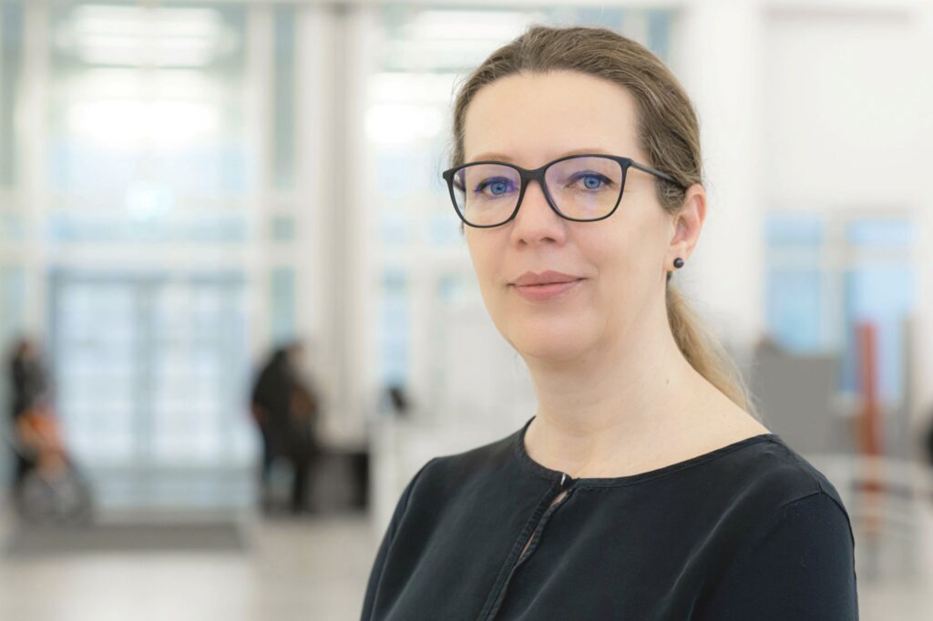 Porträtaufnahme einer Frau mit heller Haut und zurückgebundenen braunen Haaren. Sie trägt eine schwarze Bluse, eine dunkle Brille mit rechteckigem Rahmen und kleine dunkle Ohrstecker. Die Frau blickt ruhig und direkt in die Kamera. Der Hintergrund zeigt einen hellen, modern wirkenden Innenraum mit großen Fenstern und unscharfen Personen im Hintergrund. Die Atmosphäre wirkt professionell und freundlich.