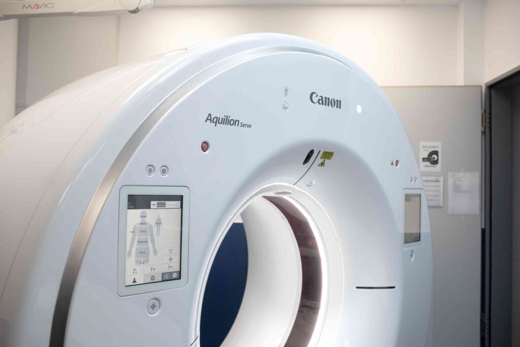 Ein moderner, weißer Computertomograph (CT-Gerät) in einem hellen Untersuchungsraum. Die große, ringförmige Öffnung des Geräts ist nach vorne ausgerichtet und innen leicht beleuchtet. Auf der Außenseite sind ein kleines Display mit einer schematischen Körpergrafik sowie Bedienelemente zu sehen. Der Markenname „Canon“ und die Modellbezeichnung „Aquilion Serve“ sind auf dem Gehäuse angebracht. Im Hintergrund stehen ein Schrank und medizinische Aushänge an der Wand.