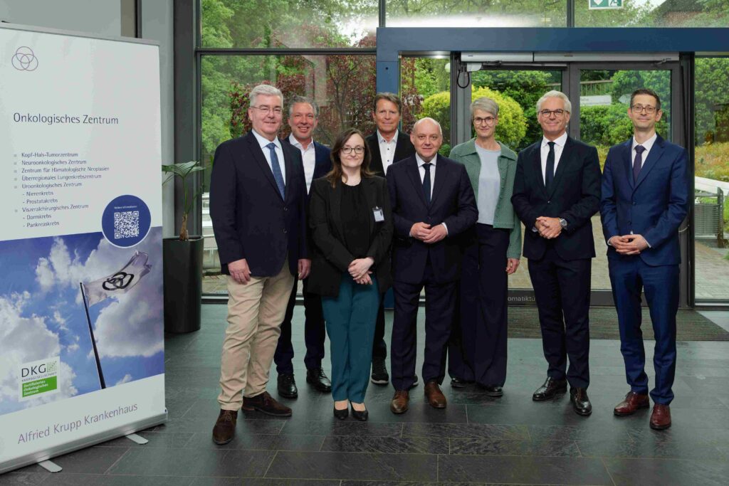 In der Eingangshalle des Alfried Krupp Krankenhauses stehen acht Personen in formeller Kleidung für ein Gruppenfoto vor dem Eingang des Onkologischen Zentrums. Links im Bild befindet sich ein großer Roll-up-Aufsteller mit der Überschrift „Onkologisches Zentrum“ sowie einer Auflistung zertifizierter Krebszentren und dem Logo der Deutschen Krebsgesellschaft. Die Gruppe blickt geschlossen in die Kamera. Im Hintergrund sind große Glasflächen mit Blick ins Grüne zu sehen. Die Atmosphäre wirkt professionell und repräsentativ.