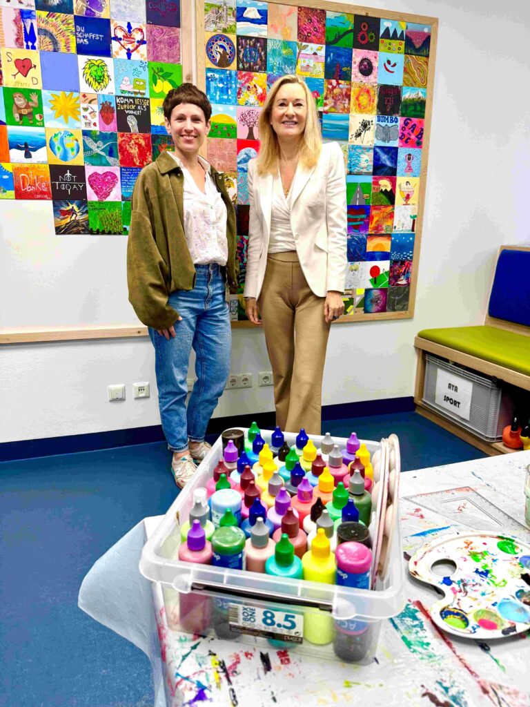 Zwei Frauen stehen in einem hellen Kreativ- und Therapieraum vor einer großen bunten Wand aus vielen kleinen quadratischen Gemälden. Die Kunstwerke zeigen unterschiedliche Motive wie Herzen, Landschaften, Blumen, abstrakte Muster und ermutigende Botschaften in kräftigen Farben. Im Vordergrund steht auf einem bemalten Arbeitstisch eine transparente Box mit zahlreichen Farbtuben und Malutensilien. Links trägt eine Frau Jeans, weiße Bluse und eine grüne Jacke, rechts steht eine blonde Frau in hellem Blazer und beigefarbener Hose. Der Raum vermittelt eine kreative, offene und freundliche Atmosphäre.