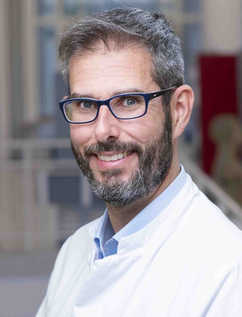 Porträtaufnahme eines lächelnden Mannes mit kurzen grau-braunen Haaren, Vollbart und dunkelblauer Brille. Er trägt einen weißen Arztkittel über einem hellblauen Hemd und blickt direkt in die Kamera. Der Hintergrund ist unscharf und zeigt eine helle klinische oder medizinische Umgebung. Die Aufnahme wirkt freundlich, professionell und vertrauensvoll.