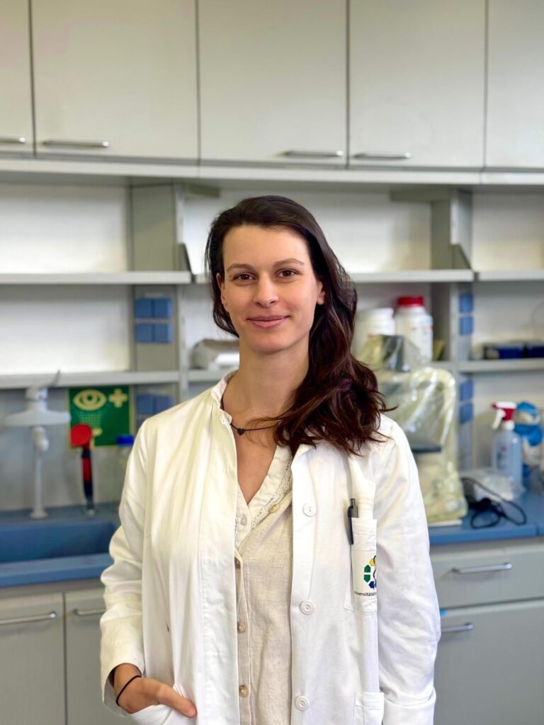 Porträt einer jungen Wissenschaftlerin in einem modernen Labor. Die Frau mit langen dunkelbraunen Haaren trägt einen weißen Laborkittel über einer hellen Bluse und blickt mit leichtem Lächeln direkt in die Kamera. Sie steht vor einer Laborarbeitsfläche mit Schränken, Geräten und Laborutensilien im Hintergrund. Auf der Arbeitsplatte sind unter anderem Sprühflaschen, Behälter und Schutzmaterialien zu sehen. Die Atmosphäre wirkt hell, sachlich und professionell und vermittelt den Eindruck biomedizinischer oder naturwissenschaftlicher Forschung.