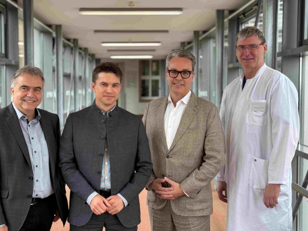 Vier Männer stehen nebeneinander auf einem hellen, verglasten Verbindungsgang eines Krankenhauses und blicken lächelnd in die Kamera. Von links nach rechts: ein Mann im dunklen Anzug mit hellblauem Hemd, ein Mann in einer dunkelgrauen Jacke mit Stehkragen, ein Mann im beigefarben karierten Anzug mit weißem Hemd sowie ein groß gewachsener Arzt im weißen Kittel. Im Hintergrund erstreckt sich der lange Flur mit großen Fensterfronten und Deckenleuchten, wodurch eine ruhige, moderne Krankenhausatmosphäre entsteht.