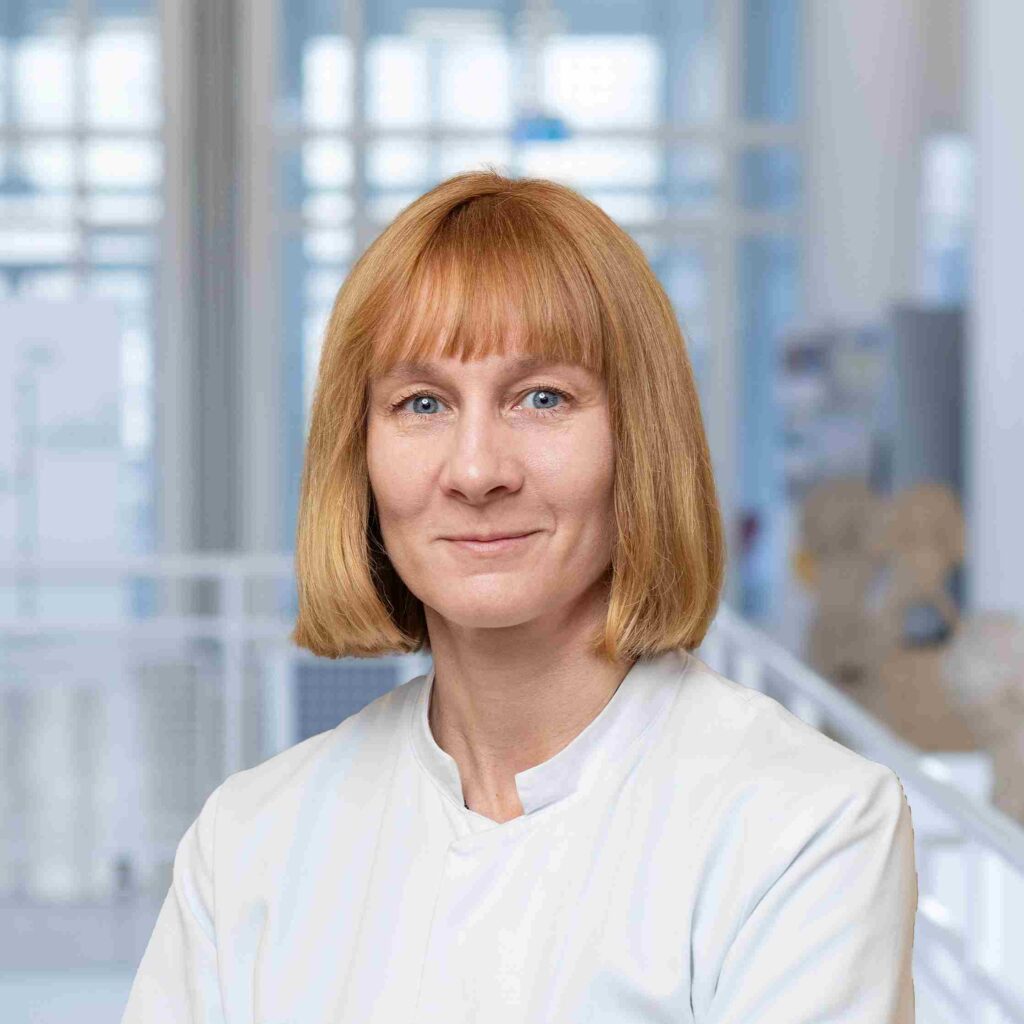 Eine Frau in weißer medizinischer Arbeitskleidung blickt direkt in die Kamera. Sie hat schulterlanges, hellblondes Haar mit geradem Pony, helle Augen und einen freundlichen, ruhigen Gesichtsausdruck. Das Porträt zeigt sie von den Schultern aufwärts vor einem unscharfen Hintergrund, der an einen hellen Klinik- oder Laborbereich erinnert. Im Hintergrund sind große Fensterflächen sowie verschwommen erkennbare medizinische Modelle oder Ausstattungsgegenstände zu sehen. Die Aufnahme wirkt professionell und vermittelt einen medizinisch-wissenschaftlichen Kontext.