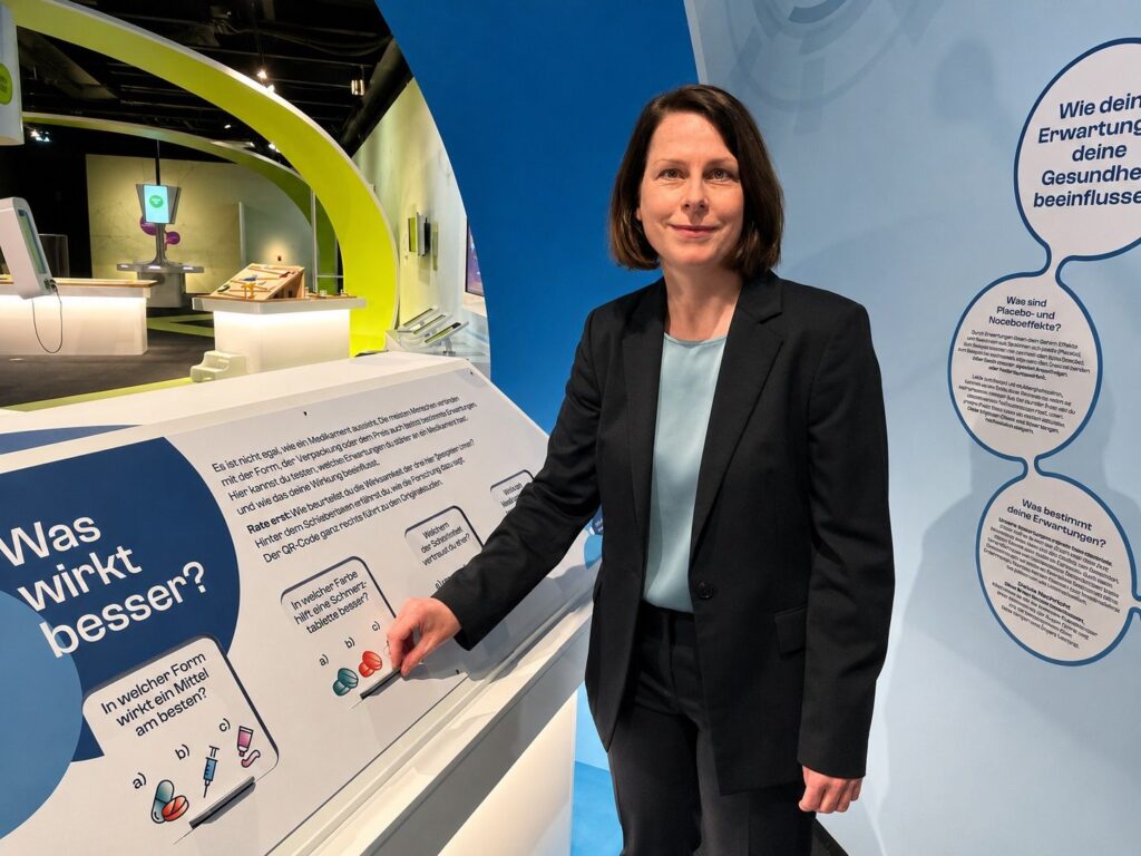 Eine Frau mit schulterlangem dunklem Haar steht in einer modernen Ausstellung und blickt direkt in die Kamera. Sie trägt einen schwarzen Hosenanzug mit hellblauer Bluse und hält einen Stift auf eine interaktive Station gerichtet. Auf der großen Tafel sind Fragen zur Wirkung von Medikamenten und zum Placebo-Effekt dargestellt, darunter „Was wirkt besser?“ sowie Abbildungen von Tabletten, Spritzen und anderen Darreichungsformen. Im Hintergrund sind weitere interaktive Exponate und Informationstafeln einer wissenschaftlichen Ausstellung zu sehen. Die hellen Blau-, Weiß- und Grüntöne verleihen dem Raum eine moderne, forschungsorientierte Atmosphäre.