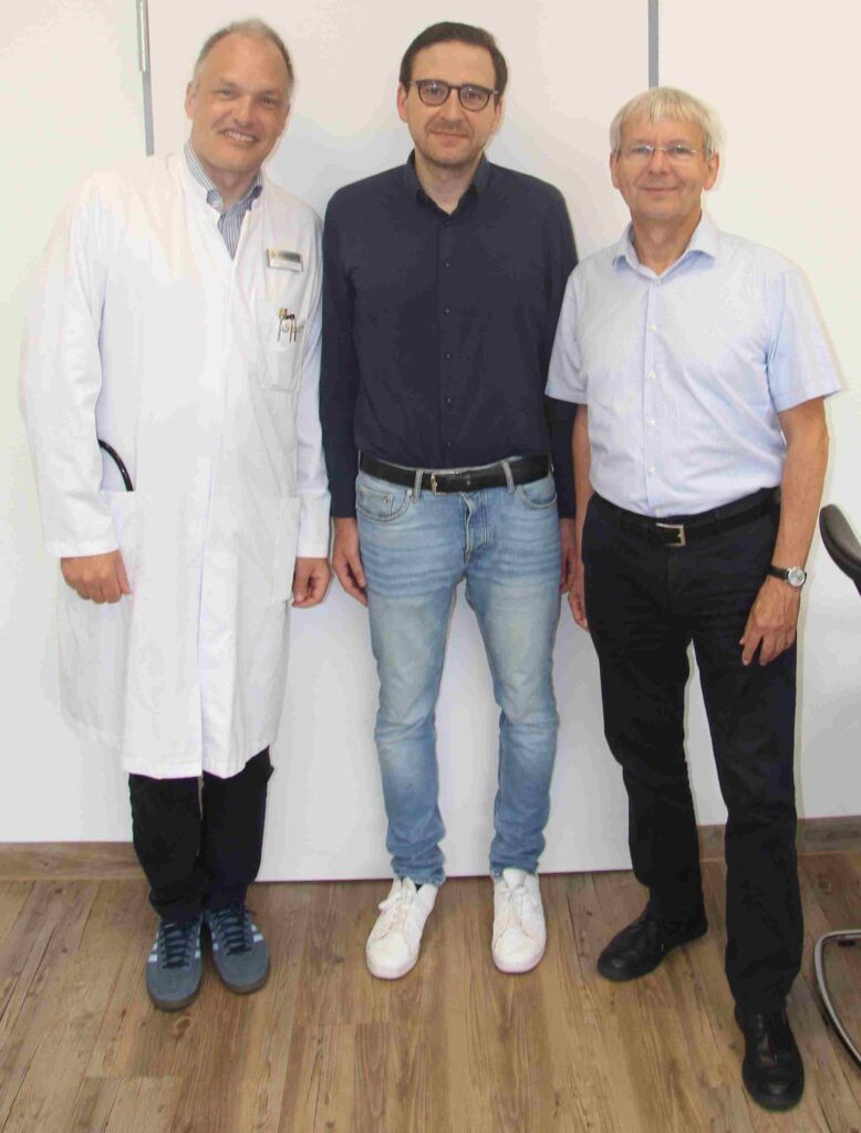 Drei Männer stehen nebeneinander in einem hellen Innenraum vor einer weißen Wand und blicken in die Kamera. Links steht ein Mann in einem weißen Arztkittel über Hemd und dunkler Hose; aus der Brusttasche ragen mehrere Stifte. In der Mitte steht ein Mann mit dunklem Hemd, heller Jeans, weißem Gürtel und weißen Turnschuhen. Er trägt eine Brille mit dunklem Rahmen. Rechts steht ein Mann mit hellblond-grauem Haar, hellblauem Kurzarmhemd, dunkler Hose und schwarzem Gürtel. Alle drei lächeln leicht. Der Boden besteht aus Holz, am rechten Bildrand ist ein Teil eines Bürostuhls zu erkennen. Die Aufnahme wirkt wie ein Gruppen- oder Erinnerungsfoto in einem klinischen oder wissenschaftlichen Umfeld.