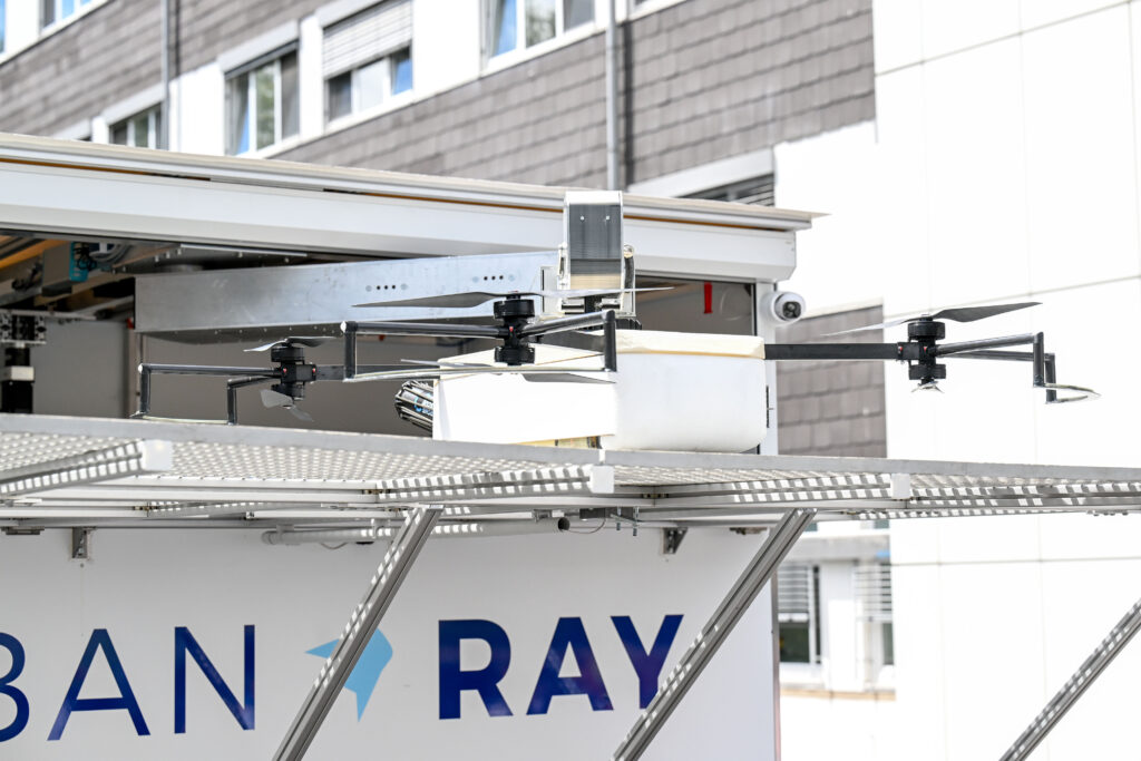 Eine kompakte Transportdrohne steht auf einer erhöhten Metallplattform vor einem mehrstöckigen Klinik- oder Institutsgebäude. Die Drohne besitzt vier Ausleger mit jeweils einem Rotor und einen länglichen, weißen Transportbehälter in der Mitte. Im Hintergrund sind Gebäudefassaden mit Fenstern sowie Teile einer technischen Infrastruktur zu erkennen. Die Aufnahme zeigt die Drohne unmittelbar vor oder nach einem Startvorgang und vermittelt den Eindruck eines Praxistests für automatisierte Transportlogistik.
