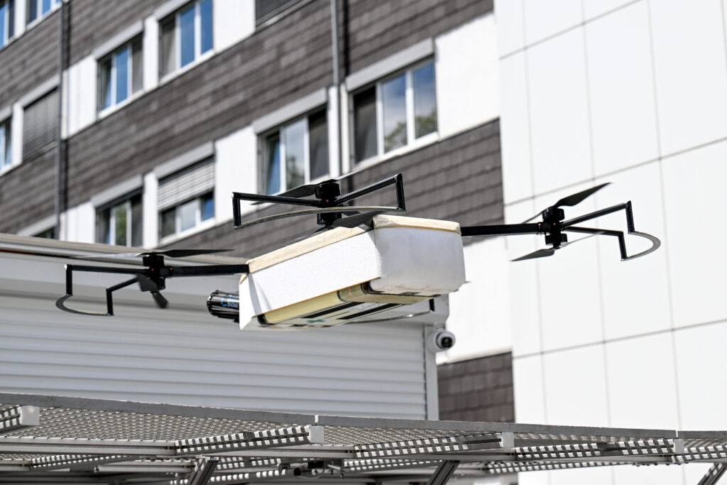 Eine weiße Transportdrohne mit vier Rotoren schwebt wenige Meter über einer Metallplattform vor einem modernen Gebäude. Der zentrale Transportbehälter ist rechteckig und an den Ecken leicht abgerundet. Die Rotoren befinden sich an schlanken Auslegern mit Schutzbügeln. Die Aufnahme dokumentiert einen Flugtest und hebt die technische Konstruktion der Drohne sowie ihre Eignung für den Transport kleiner Güter hervor.