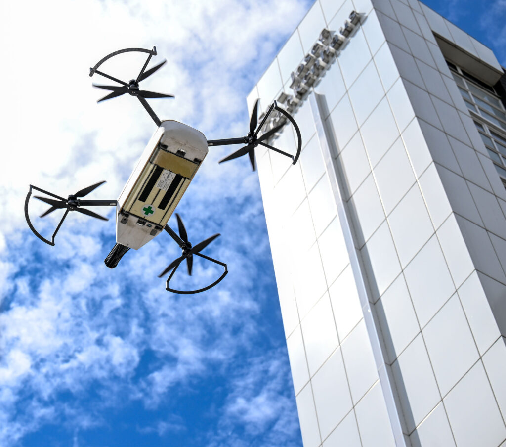 Aus einer Untersicht fotografiert fliegt eine Transportdrohne vor einem hohen, hellen Klinik- oder Institutsgebäude. Die vier Rotoren sind deutlich sichtbar, ebenso die Unterseite des Transportbehälters mit einem grünen Kreuz als Hinweis auf medizinische Nutzung. Über der Drohne erstreckt sich ein teilweise bewölkter blauer Himmel. Das Bild vermittelt den Einsatz moderner Drohnentechnologie für medizinische oder logistische Transportaufgaben im Gesundheitswesen.