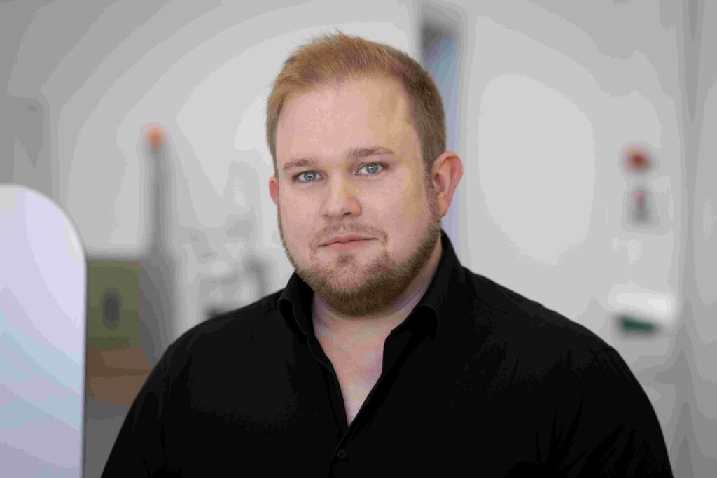 Porträt eines Mannes mit kurzem blondem Haar, hellem Bart und blauen Augen. Er blickt direkt in die Kamera und trägt ein schwarzes Hemd mit offenem Kragen. Das Foto zeigt ihn von der Brust aufwärts vor einem unscharfen Hintergrund in hellen Grau- und Weißtönen, der an ein modernes Büro- oder Forschungsumfeld erinnert. Die Person ist zentral im Bild positioniert, die Beleuchtung ist gleichmäßig und professionell. Der Gesamteindruck wirkt sachlich, kompetent und freundlich.