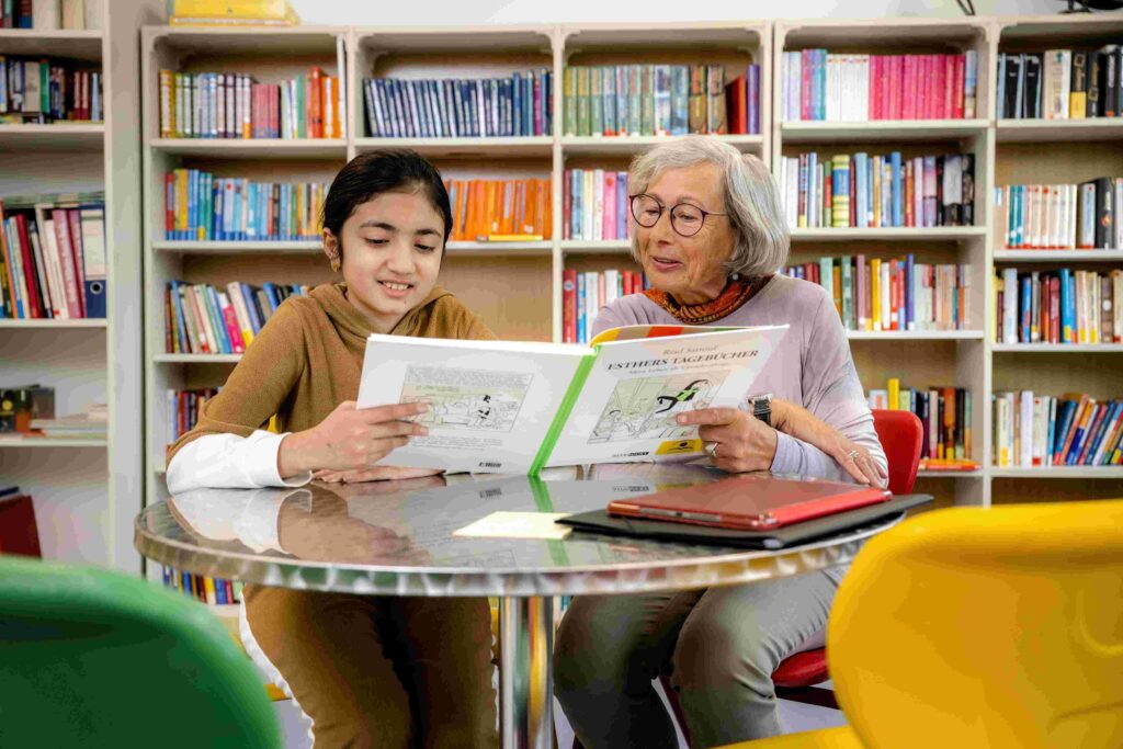 In einer Bibliothek sitzen ein Kind und eine ältere Frau gemeinsam an einem runden Tisch und lesen in einem aufgeschlagenen Buch. Beide blicken aufmerksam auf die Seiten und wirken konzentriert und interessiert. Hinter ihnen stehen hohe Bücherregale mit zahlreichen farbigen Büchern. Auf dem Tisch liegen weitere Unterlagen sowie ein Tablet in einer roten Schutzhülle. Die Szene vermittelt eine ruhige Lern- und Vorlesesituation zwischen den Generationen in einer freundlichen, hellen Umgebung.