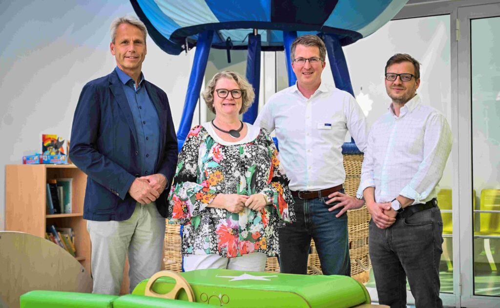 Vier Personen stehen nebeneinander in einem hellen, freundlich gestalteten Innenraum und blicken lächelnd in die Kamera. Im Hintergrund befindet sich ein großer dekorativer Heißluftballonkorb in Blau und Weiß sowie eine Spielecke mit Regal und Kinderbüchern, was auf eine kinderfreundliche Umgebung hinweist. Die Personen tragen überwiegend Business-Casual-Kleidung. Im Vordergrund ist ein grünes Spiel- oder Bewegungsmodul zu sehen.