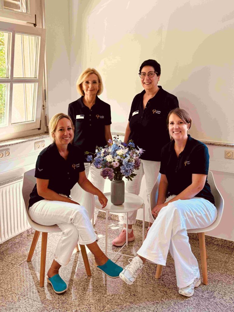 Vier Mitarbeitende eines medizinischen Teams posieren in einem hellen Praxisraum für ein Gruppenfoto. Zwei Frauen sitzen auf hellen Holzstühlen, zwei weitere stehen dahinter. Alle tragen dunkelblaue Poloshirts mit Logo und weiße Hosen. Zwischen ihnen steht auf einem kleinen runden Beistelltisch eine graue Vase mit einem blau-weiß-lila Blumenstrauß. Links fällt Tageslicht durch ein geöffnetes Fenster in den Raum.