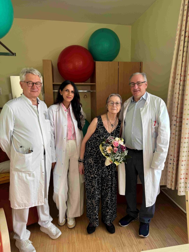 Vier Personen stehen in einem Therapieraum einer Klinik nebeneinander. Zwei Ärzte und eine Ärztin begleiten eine ältere Frau, die einen Blumenstrauß in den Händen hält. Alle blicken lächelnd in die Kamera.
