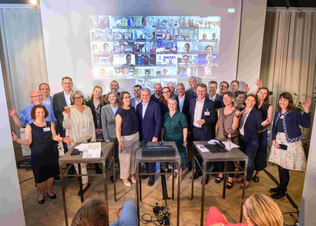 Gruppenfoto einer wissenschaftlichen Veranstaltung mit rund 30 Teilnehmenden auf einer Bühne. Die Personen stehen lächelnd nebeneinander und blicken in die Kamera, einige winken. Im Hintergrund ist auf einer großen Leinwand eine Videokonferenz mit zahlreichen zugeschalteten Teilnehmenden zu sehen. Vor der Gruppe stehen drei Stehtische mit Präsentationstechnik.
