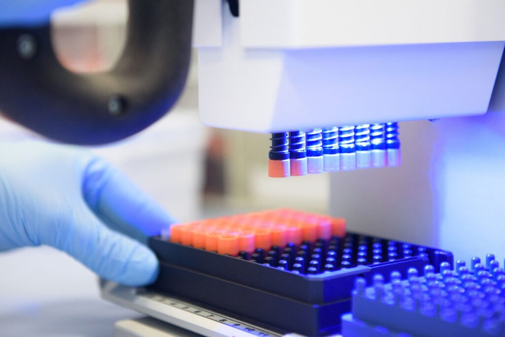 Nahaufnahme eines automatisierten Laborgeräts, das mehrere Probenröhrchen über einer Mikrotiterplatte positioniert. Eine behandschuhte Hand schiebt den Probenträger in das Gerät. Blaues Arbeitslicht beleuchtet die Laborumgebung und unterstreicht den technischen Charakter der Aufnahme