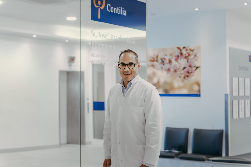 Ein Arzt in weißem Kittel steht lächelnd im hellen Eingangsbereich des St. Josef-Krankenhauses Kupferdreh. Hinter ihm sind das Contilia-Logo auf einem Deckenbanner, Sitzgelegenheiten sowie ein großformatiges Bild mit Blütenmotiv zu sehen. Die Aufnahme vermittelt eine freundliche und moderne Krankenhausatmosphäre.