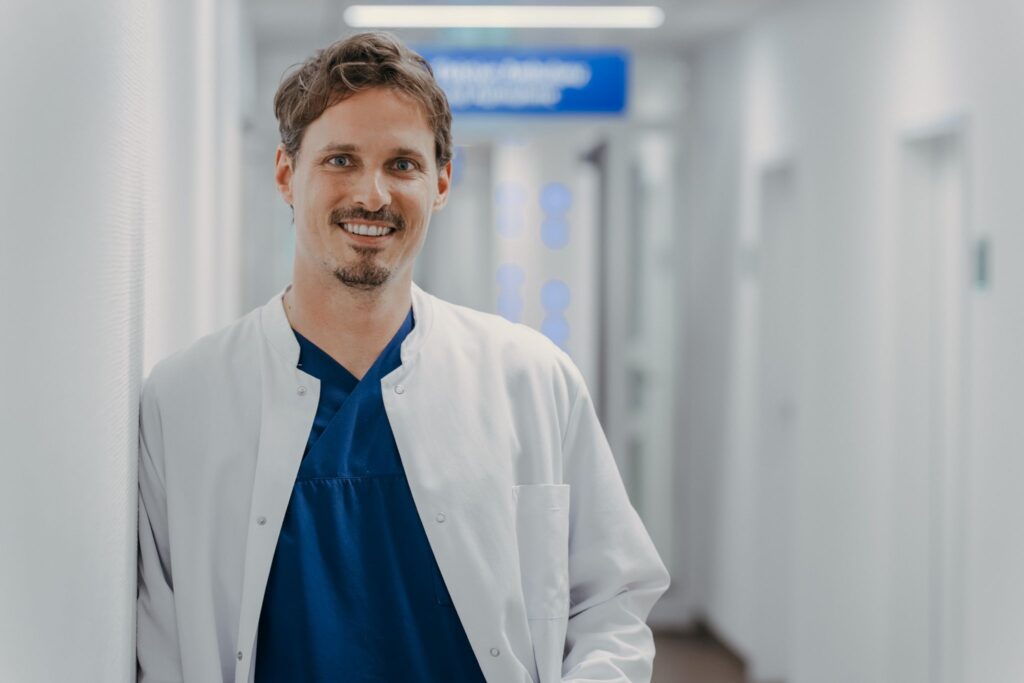 Ein lächelnder Arzt steht in einem hellen Krankenhausflur und blickt direkt in die Kamera. Er trägt einen weißen Arztkittel über dunkelblauer OP-Kleidung und lehnt entspannt an einer Wand. Im unscharfen Hintergrund sind Türen sowie ein blaues Hinweisschild des Klinikflurs zu erkennen.