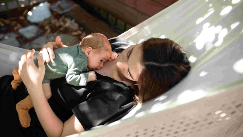 Eine Mutter liegt entspannt in einer Hängematte und hält ihr Neugeborenes eng an ihrer Brust. Das Baby liegt in Bauchlage im Hautkontakt auf der Mutter und blickt aufmerksam nach vorne. Sonnenlicht fällt durch Blätter und erzeugt helle Lichtflecken auf der Hängematte und der Kleidung. Die ruhige, geschützte Atmosphäre vermittelt Nähe, Geborgenheit und die frühe Bindung zwischen Mutter und Kind.