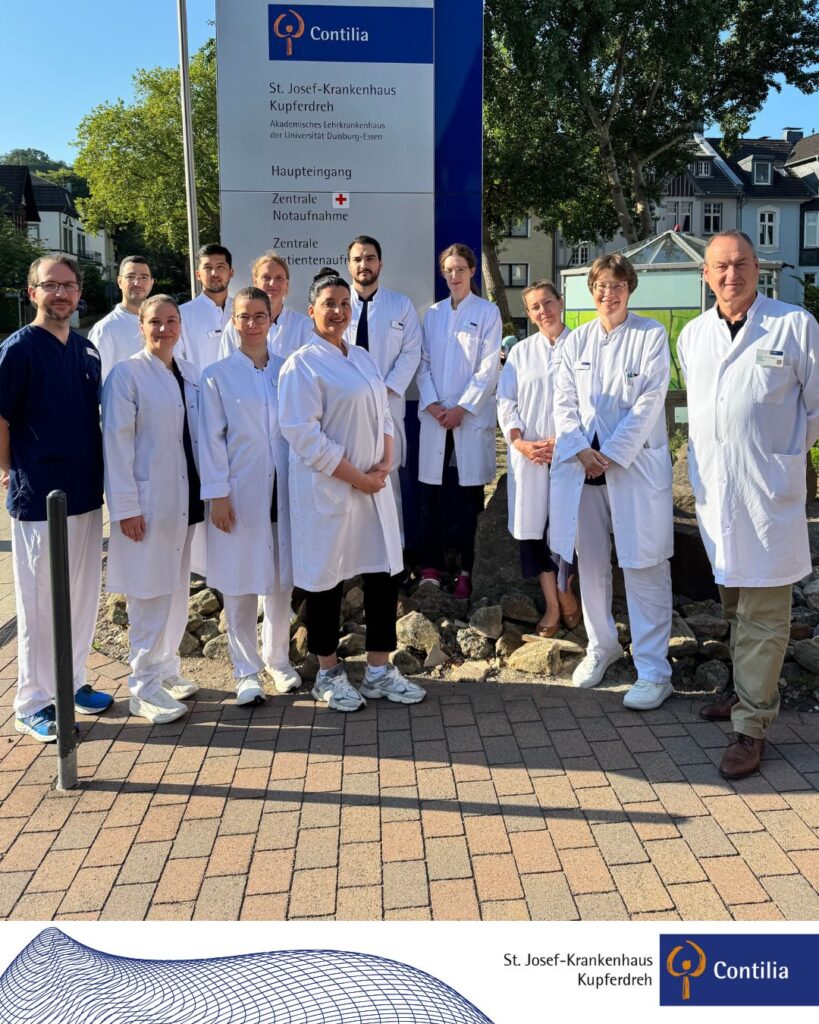 Eine Gruppe von elf Mitarbeitenden des St. Josef-Krankenhauses Kupferdreh steht in weißen Arzt- und Pflegekitteln vor dem Haupteingang des Krankenhauses. Die Personen sind halbkreisförmig vor einem großen Wegweiser mit der Aufschrift „St. Josef-Krankenhaus Kupferdreh“ angeordnet und blicken lächelnd in die Kamera. Im Hintergrund sind Bäume sowie Wohnhäuser zu sehen. Das Foto entstand bei sonnigem Wetter im Außenbereich der Klinik.