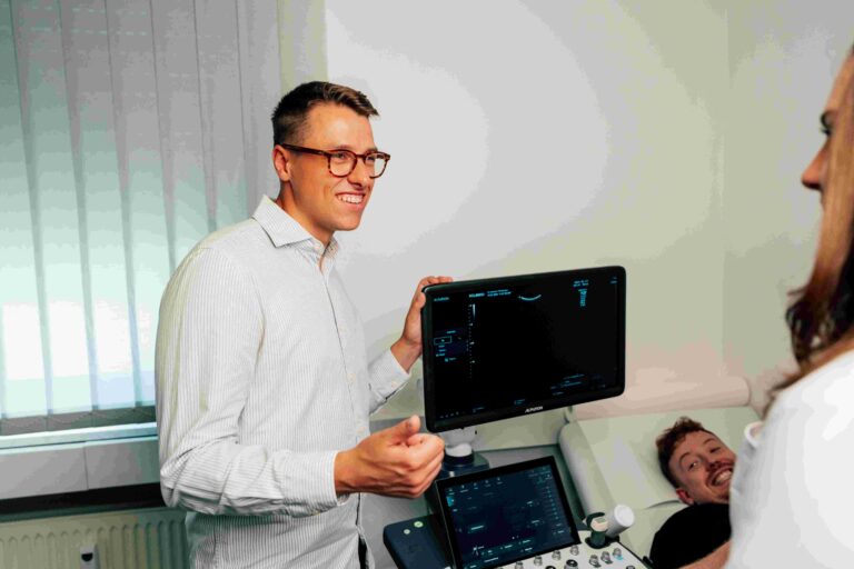 Drei Personen in einem hellen Untersuchungsraum an einem Ultraschallgerät. Ein lächelnder Mann mit Brille steht am Gerät und spricht mit einer Frau im Vordergrund. Rechts liegt ein weiterer Mann auf einer Untersuchungsliege und blickt lächelnd zu ihnen.