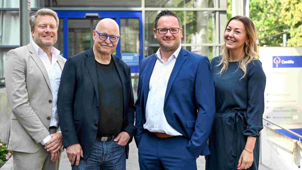 Vier Personen stehen lächelnd nebeneinander vor dem Eingangsbereich eines Gebäudes. Drei Männer und eine Frau blicken in die Kamera. Im Hintergrund sind große Glasflächen, ein blau gerahmter Eingang sowie ein Wegweiser mit dem Contilia-Logo zu sehen.
