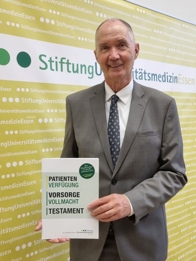 Ein lächelnder älterer Mann im grauen Anzug hält vor einer grün-weißen Wand der Stiftung Universitätsmedizin Essen einen Vorsorgeratgeber in die Kamera. Auf dem Titel stehen groß die Themen „Patientenverfügung“, „Vorsorgevollmacht“ und „Testament“.