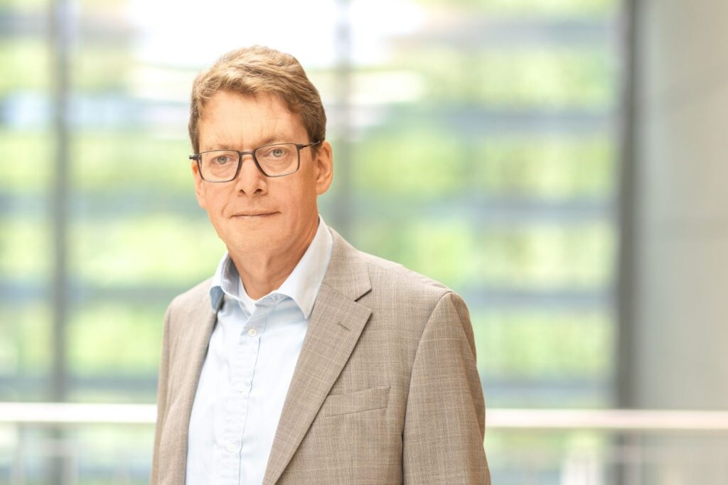 Porträt eines Mannes mit kurzen braunen Haaren und Brille. Er trägt ein hellblaues Hemd und ein hellgraues Sakko und blickt in die Kamera. Der Hintergrund ist unscharf.