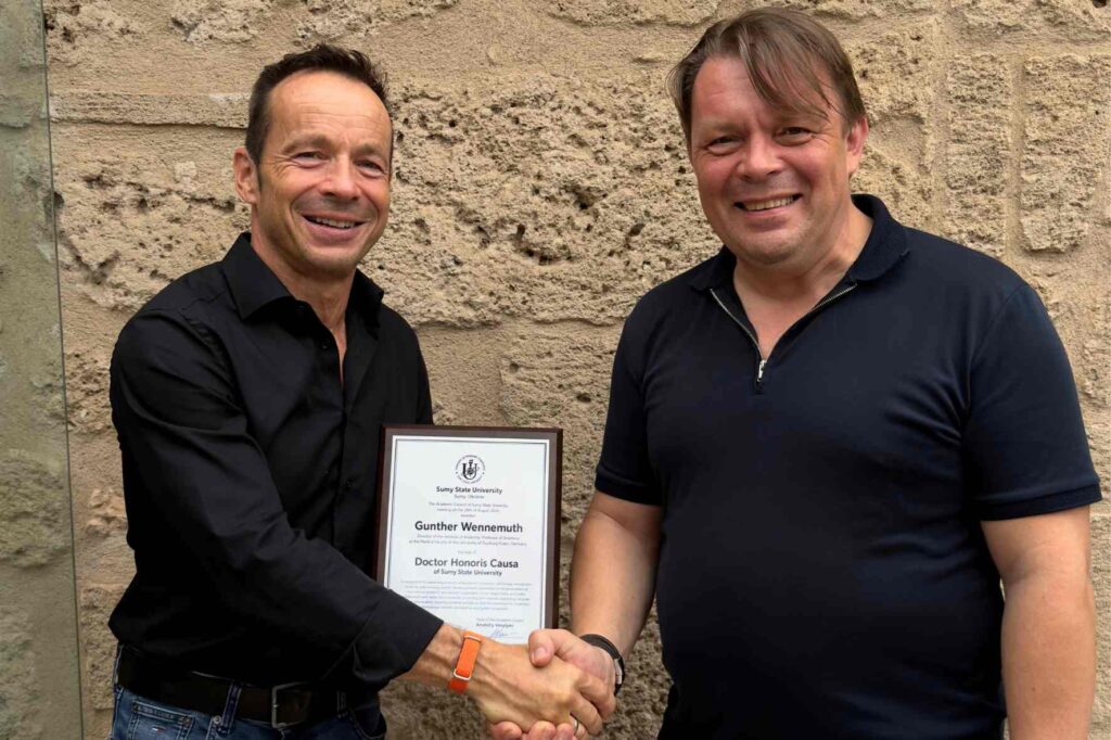 Zwei lächelnde Männer stehen vor einer hellen Steinwand und geben sich die Hand. Der Mann links hält eine gerahmte Urkunde zur Verleihung eines Ehrendoktortitels der Sumy State University.