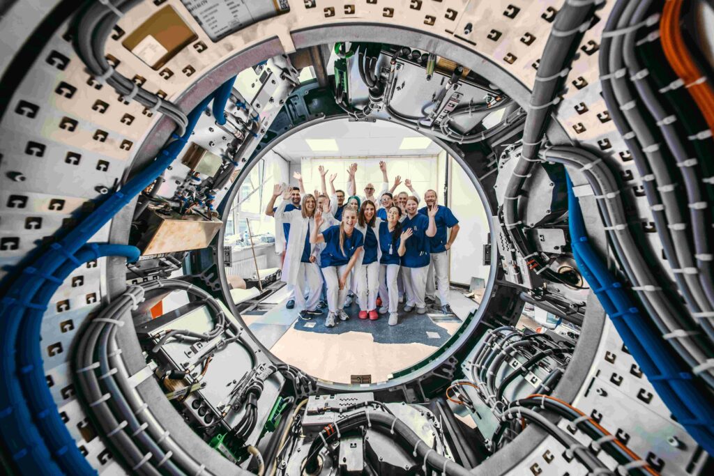 Blick durch die runde Öffnung eines CT-Geräts mit zahlreichen Kabeln und technischen Bauteilen. Dahinter steht eine Gruppe von Mitarbeitenden in medizinischer Arbeitskleidung und weißen Kitteln, die lächelnd in die Kamera schaut und teilweise die Hände hebt.
