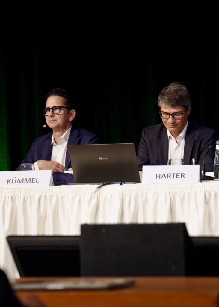 Zwei Männer sitzen bei einer Fachveranstaltung nebeneinander an einem Podiumstisch. Vor ihnen stehen Namensschilder mit den Aufschriften „Kümmel“ und „Harter“. Zwischen ihnen befindet sich ein geöffneter Laptop. Beide tragen dunkle Anzüge, weiße Hemden und Brillen; der Mann links trägt ein Headset-Mikrofon. Im Hintergrund ist eine dunkel gehaltene Bühne mit grüner Beleuchtung zu sehen.