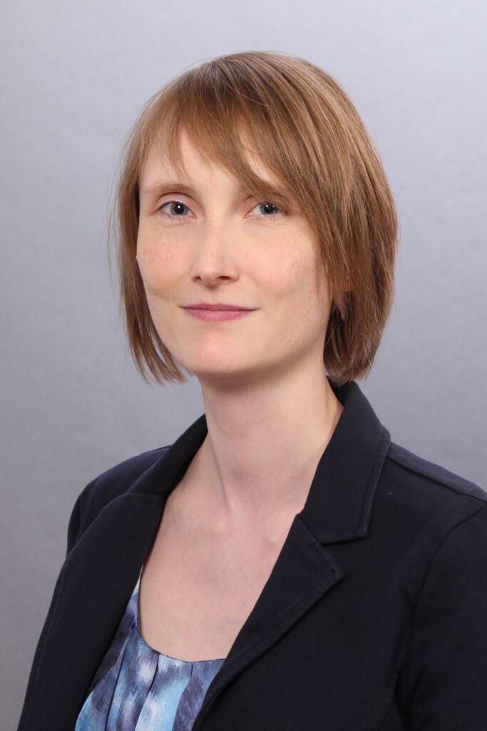 Porträt einer Frau mit kurzem, hellbraunem Haar vor einem neutralen grauen Hintergrund. Sie trägt einen dunklen Blazer über einem blau gemusterten Oberteil und blickt mit leichtem Lächeln in die Kamera.