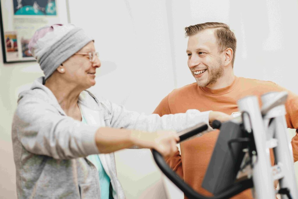 Eine Frau mit Kopfbedeckung trainiert an einem medizinischen Trainingsgerät. Neben ihr sitzt ein lächelnder Mann, der sie während der Übung begleitet.