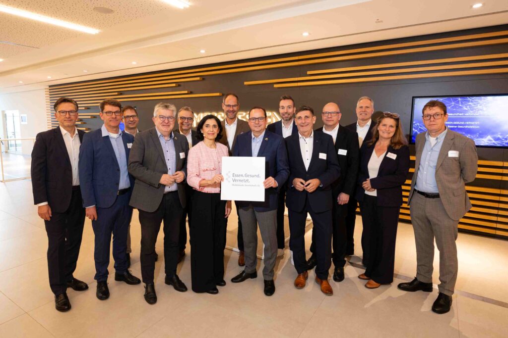Gruppenfoto von 13 Personen, darunter zwei Frauen und elf Männer, in einem modernen Veranstaltungsraum. Die Gruppe steht in zwei Reihen vor einer dunklen Wand mit horizontalen Holzleisten. Im Mittelpunkt halten eine Frau und ein Mann ein weißes Schild mit dem Logo von „Essen.Gesund.Vernetzt. – Medizinische Gesellschaft e. V.“. Rechts im Hintergrund zeigt ein Bildschirm das Motiv des 11. Essener Gesundheitsforums. Alle Personen blicken in die Kamera.