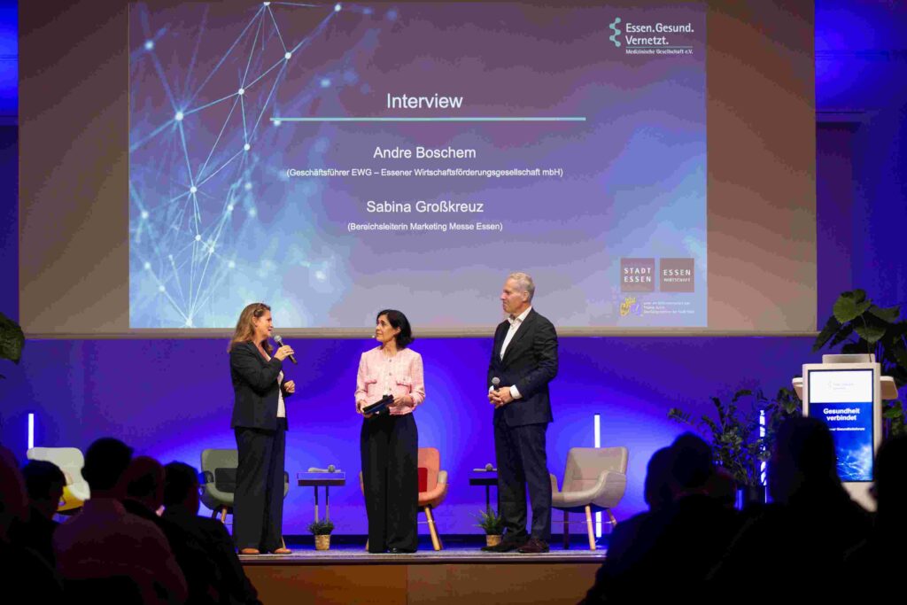 Drei Personen stehen auf der blau beleuchteten Bühne des 11. Essener Gesundheitsforums. Links spricht Sabina Großkreuz, Bereichsleiterin Marketing der Messe Essen, in ein Mikrofon. In der Mitte steht die Moderatorin mit Moderationskarten, rechts Andre Boschem, Geschäftsführer der Essener Wirtschaftsförderung, mit einem Mikrofon. Im Hintergrund zeigt eine große Leinwand die Namen und Funktionen der beiden Interviewgäste. Im Vordergrund sind die Silhouetten des Publikums zu sehen.