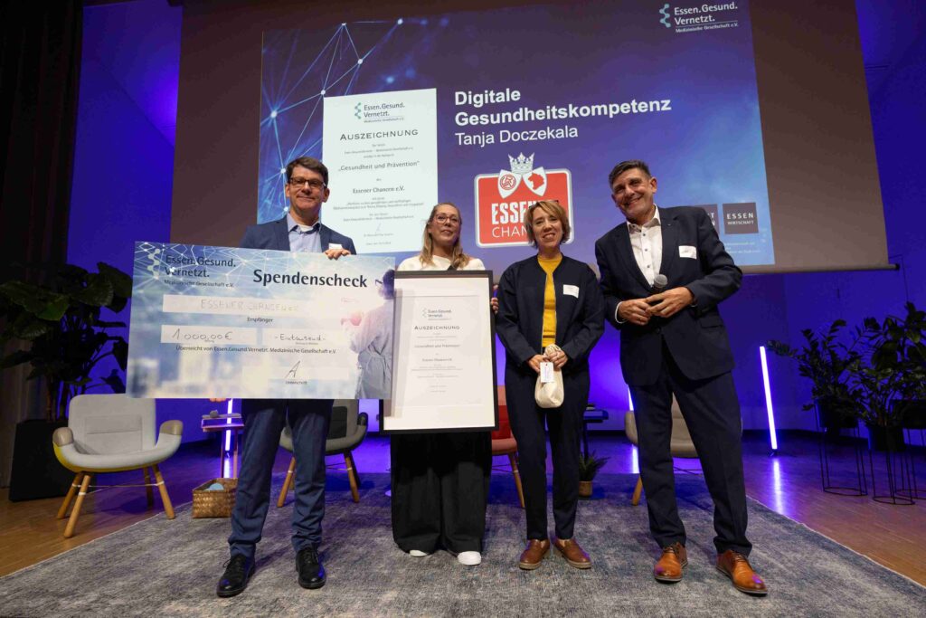 Vier Personen, darunter drei Frauen und ein Mann, stehen bei der Verleihung des Jahrespreises „Gesundheit und Prävention“ auf der blau beleuchteten Bühne des 11. Essener Gesundheitsforums. Sie präsentieren eine gerahmte Urkunde und einen großen Spendenscheck über 1.000 Euro. Ausgezeichnet wird der Verein Essener Chancen e. V. für sein Projekt zur digitalen Gesundheitskompetenz. Im Hintergrund zeigt eine große Leinwand den Projekttitel, den Namen Tanja Doczekala und das Logo von Essener Chancen.